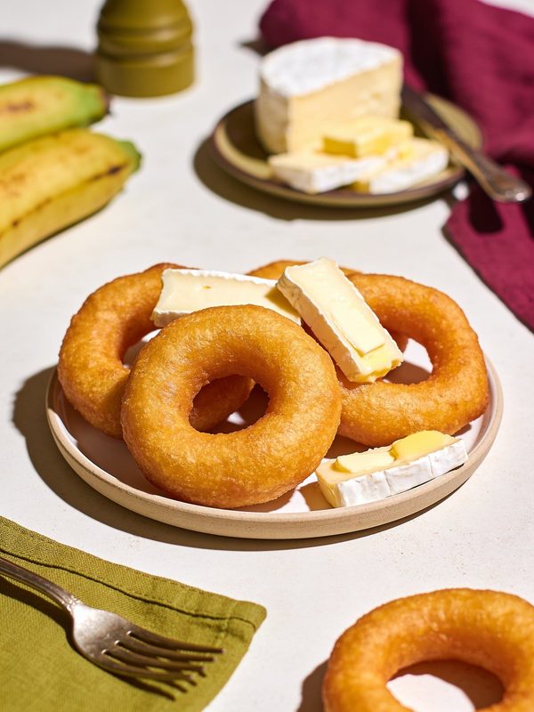 A plate of golden fried mandoca rings with slices of soft white cheese, ripe plantains in the background.