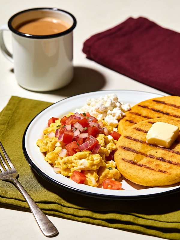 Venezuelan perico topped with diced tomato and onion, with grilled arepas, butter, crumbled white cheese and a cup of coffee.